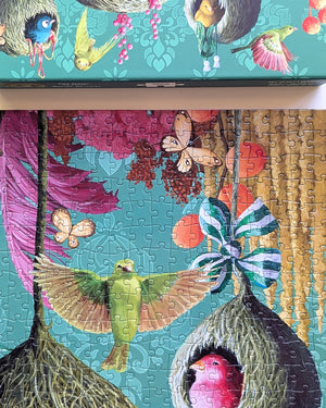 A completed jigsaw puzzle showing a colorful nature scene with birds, butterflies, and nests on a turquoise background, with part of the matching puzzle box visible at the top.