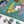 A close-up of a completed jigsaw puzzle showing a colorful scene with a yellow bird, blue bird, green patterned background, and pink flowers, with several loose puzzle pieces placed on a white surface below.