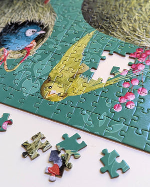 A close-up of a completed jigsaw puzzle showing a colorful scene with a yellow bird, blue bird, green patterned background, and pink flowers, with several loose puzzle pieces placed on a white surface below.