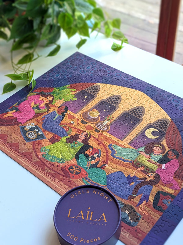 Completed puzzle of women sitting in a large open room enjoying each others company. Puzzle is on a white desk surrounded by plants and box top lid from puzzle. 