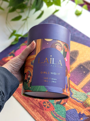 Hand holding a Laila Luxury puzzle box for the puzzle 'Girls Night' shown over portion of the completed puzzle on a white desk with plants.