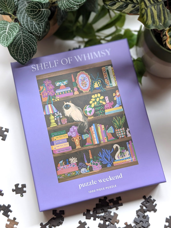 Jigsaw puzzle box titled 'Shelf of Whimsy' with a cat illustration on a white surface.