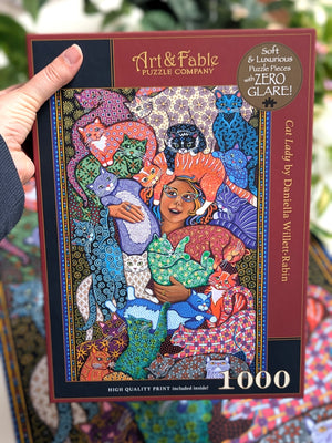 Woman's hand holding Art & Fable puzzle box showing an illustration woman holding many different patterned cats. 