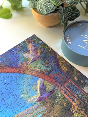 Close up of corner of the completed Golden Domes puzzle showing vibrant archway and flying pigeons. Next to plant and top of tube packaging.