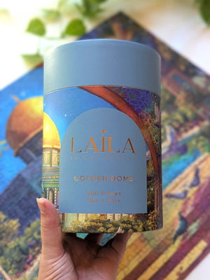 Hand holding a puzzle packaged in a tube style container in front of a completed version of the puzzle blurred in the background