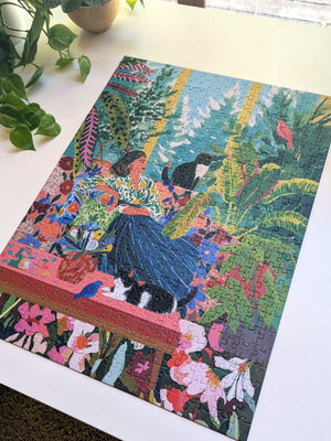 A completed colorful jigsaw puzzle featuring a woman in a blue skirt among tropical plants and flowers, displayed on a white tabletop with a plant nearby.