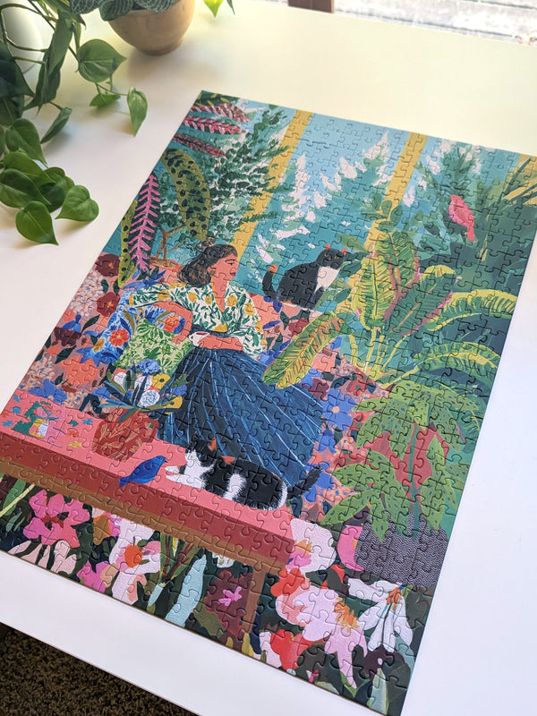 A completed colorful jigsaw puzzle featuring a woman in a blue skirt among tropical plants and flowers, displayed on a white tabletop with a plant nearby.