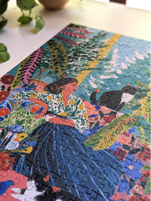 A colorful completed jigsaw puzzle featuring a stylized illustration of a woman in a floral blouse and dark skirt, surrounded by tropical leaves and cats, photographed on a light tabletop with a plant leaf in the corner.