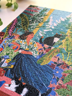 A colorful, fully assembled jigsaw puzzle featuring a vibrant illustration of a woman in a blue skirt surrounded by tropical plants and two black-and-white cats, photographed on a light surface with a small plant leaf in the corner.