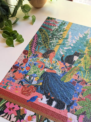 A colorful, fully assembled jigsaw puzzle featuring a whimsical painting of a woman in a blue dress with a black and white cat, displayed on a light-colored tabletop next to a potted plant.