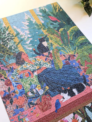 A completed jigsaw puzzle featuring a colorful tropical scene with a woman, cats, birds, and lush green foliage, photographed on a light surface.