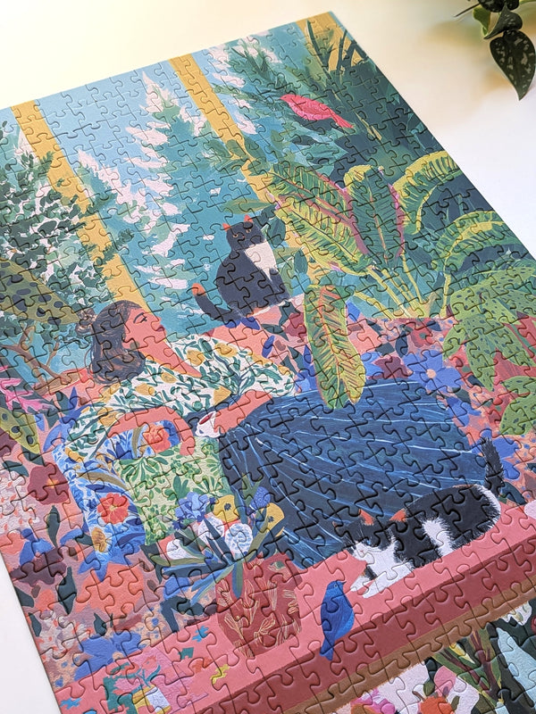 A completed jigsaw puzzle featuring a colorful tropical scene with a woman, cats, birds, and lush green foliage, photographed on a light surface.
