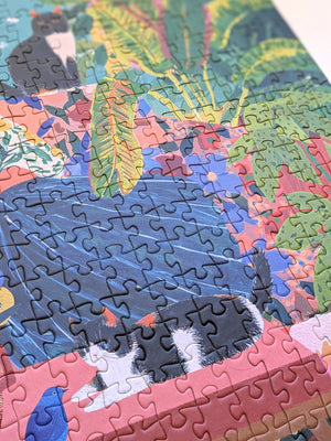 A colorful completed jigsaw puzzle showing a tropical-style scene with a black-and-white cat, blue water, green leaves, and pink foreground elements.