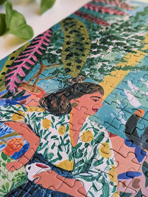 A colorful jigsaw puzzle featuring a stylized portrait of a woman with tropical plants, shown in a close-up angled shot with a white surface and green leaves in the background.
