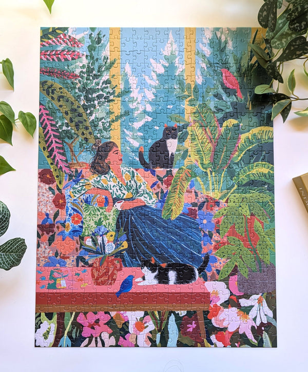 A completed jigsaw puzzle featuring a colorful, detailed illustration of a woman in a lush garden with cats and flowers, displayed on a white surface with decorative green leaves around it.