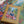 A boxed jigsaw puzzle called 'INDOOR ARBORETEM' by Puzzle Weekend is placed on top of a completed multicolor puzzle. The box is tan with a colorful illustration of a woman in a garden, and the background shows additional puzzle pieces and leaves.