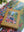 A boxed jigsaw puzzle called 'INDOOR ARBORETEM' by Puzzle Weekend is placed on top of a completed multicolor puzzle. The box is tan with a colorful illustration of a woman in a garden, and the background shows additional puzzle pieces and leaves.