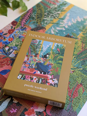 A boxed jigsaw puzzle called 'INDOOR ARBORETEM' by Puzzle Weekend is placed on top of a completed multicolor puzzle. The box is tan with a colorful illustration of a woman in a garden, and the background shows additional puzzle pieces and leaves.