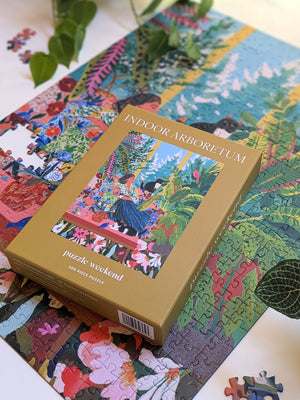 A boxed jigsaw puzzle titled 'INDOOR ARBOREtum' by puzzle weekend, placed on top of a completed multicolor botanical puzzle. The box is tan/gold with an illustrated forest scene on the front.