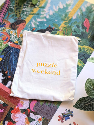 A small off-white fabric drawstring pouch with the words 'puzzle weekend' printed in yellow lowercase letters, placed on top of a colorful jigsaw puzzle.