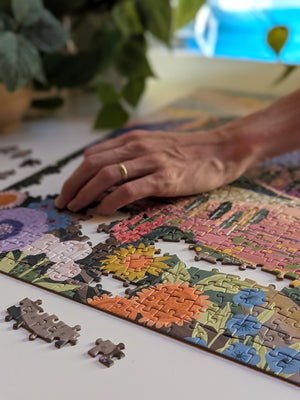 A hand sits atop a nearly complete jigsaw puzzle with floral artwork. Puzzle is on white desk near plants.