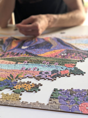 An out of focus person sits behind a white desktop upon which a partially completed mountain scene puzzle sits.