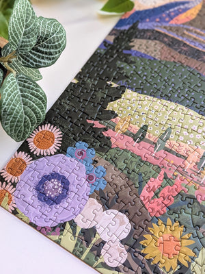 Close-up of the corner of a completed puzzle showing flowers and trees. Puzzle is on a white surface near plants.