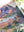 Nearly half of a completed puzzle with  mountain scene artwork is visible on a white surface near plants and the corner of a puzzle box lid.