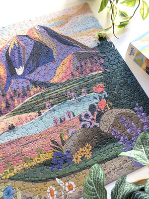 Nearly half of a completed puzzle with  mountain scene artwork is visible on a white surface near plants and the corner of a puzzle box lid.