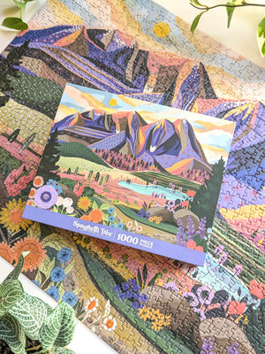 Mountain and Valley themed jigsaw puzzle box sits atop a completed copy of the puzzle on a white surface with some plants visible. 