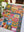A colorful patchwork jigsaw puzzle featuring whimsical folk-art designs like flowers, a rainbow, a mushroom, a butterfly, and geometric patterns, displayed on a white table with a plant in the foreground.