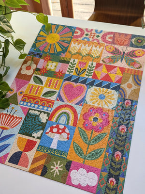 A colorful patchwork jigsaw puzzle featuring whimsical folk-art designs like flowers, a rainbow, a mushroom, a butterfly, and geometric patterns, displayed on a white table with a plant in the foreground.