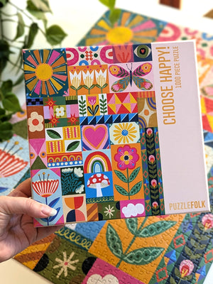 A hand holds a colorful patchwork jigsaw puzzle box labeled 'CHOOSE HAPPY! 1000 PIECE PUZZLE' by PuzzleFolk, with a matching completed jigsaw puzzle visible in the background.