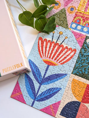 Partial view of a completed jigsaw puzzle with a graphic red and white flower with blue leaves near a puzzle box lid and plants on a white desk surface.