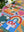 Close-up of a partial view of a completed jigsaw puzzle focusing on a red and white mushroom on a blue square and a colorful rainbow on a white desktop with plants.