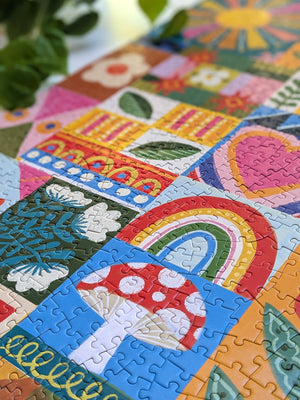 Close-up of a partial view of a completed jigsaw puzzle focusing on a red and white mushroom on a blue square and a colorful rainbow on a white desktop with plants.