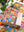 Photo of a jigsaw puzzle box with the text 'Choose Happy' and a patchwork of colorful artwork featuring flowers, patterns and butterflies on top of a completed version of the puzzle on a white desk surrounded by plants. 
