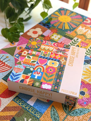 Photo of a jigsaw puzzle box with the text 'Choose Happy' and a patchwork of colorful artwork featuring flowers, patterns and butterflies on top of a completed version of the puzzle on a white desk surrounded by plants. 