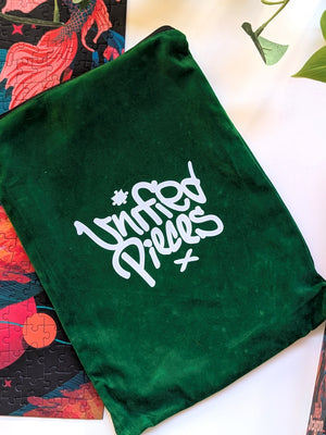 Velvet forest green zippered pouch with white Unified Pieces logo printed in the middle. Pouch sits on a partial view of a completed puzzle and green plants are shown in the top right. 