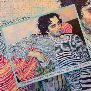 Jigsaw puzzle box showing a colorful modern portrait of the artist Yuri Masnyj and the text 'Serious Snack by Hope Gangloff'. Box is on top of a completed close up of the same puzzle.