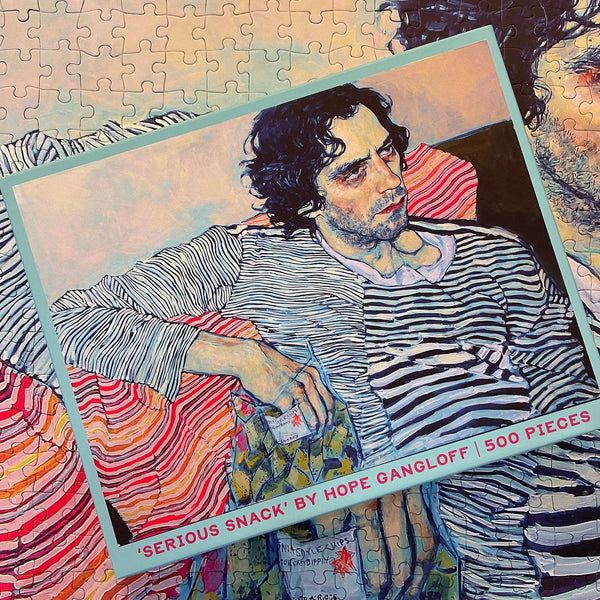 Jigsaw puzzle box showing a colorful modern portrait of the artist Yuri Masnyj and the text 'Serious Snack by Hope Gangloff'. Box is on top of a completed close up of the same puzzle.