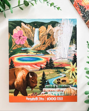 A 1000-piece jigsaw puzzle box titled 'Spaghetti Tree' featuring a colorful illustrated scene of Yellowstone National Park with a bison, waterfall, geysers, and hot springs, photographed on a white surface with decorative greenery.