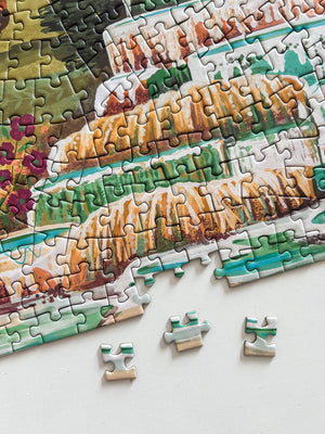 A close-up of a completed jigsaw puzzle showing a colorful landscape scene with layered tan and green hills, water, and foliage. Three loose puzzle pieces are placed on a white surface below the puzzle.