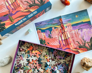 Open jigsaw puzzle box with pieces on top of a poster of the puzzle showing nighttime desert scenery. Box and poster are on white surface surrounded by box lid and stones. 