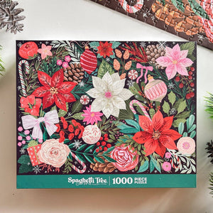 A 1000-piece jigsaw puzzle box by Spaghetti Tree, featuring a festive Christmas illustration with poinsettias, candy canes, ornaments, pinecones, and bows on a dark background. The box is displayed on a light surface with decorative pinecones and a puzzle edge visible around it.