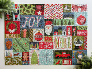 A holiday jigsaw puzzle featuring Christmas-themed blocks with the words JOY, PEACE, NOEL, and JOLLY, plus ornaments, Santa, trees, candy canes, snowflakes, and holiday motifs. Pine branches frame the top and bottom corners against a white surface.