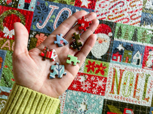 A hand holds several small, colorful Christmas-themed jigsaw puzzle pieces in front of a festive quilted background with Santa and holiday motifs.