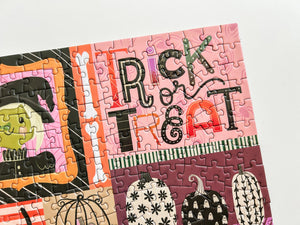 A close-up of a completed jigsaw puzzle featuring a pink and orange Halloween design with the words 'Trick or Treat' in decorative lettering, along with pumpkins, ghosts, and a witch illustration, photographed on a white background.