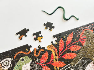 A Halloween-themed jigsaw puzzle next to a fake snake. The puzzle has several loose pieces scattered on a white surface, including a frog, spiderwebs, and orange autumn leaves on a dark background.