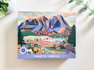 Top view of puzzle box showing illustration of beautiful mountain and valley scene. Box is on white background surrounded by small foliage.